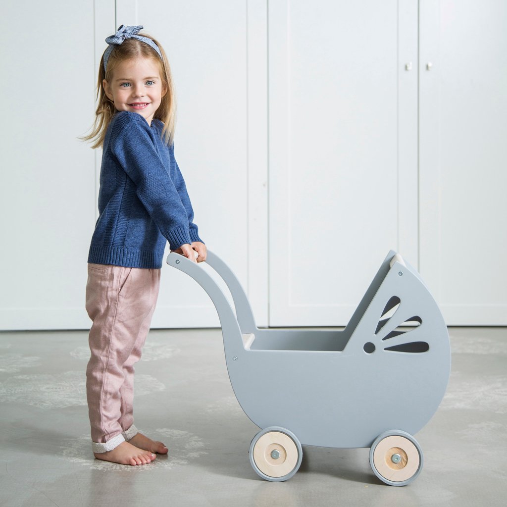 Kind schiebt Puppenwagen Lilli aus Holz im Kinderzimmer Kind schiebt im Innenraum einen grauen Puppenwagen Lilli aus Holz mit vier Rädern, im Hintergrund helle Schrankfront, Kind trägt blauen Pullover, rosa Hose und Haarband, barfuß auf grauem Boden