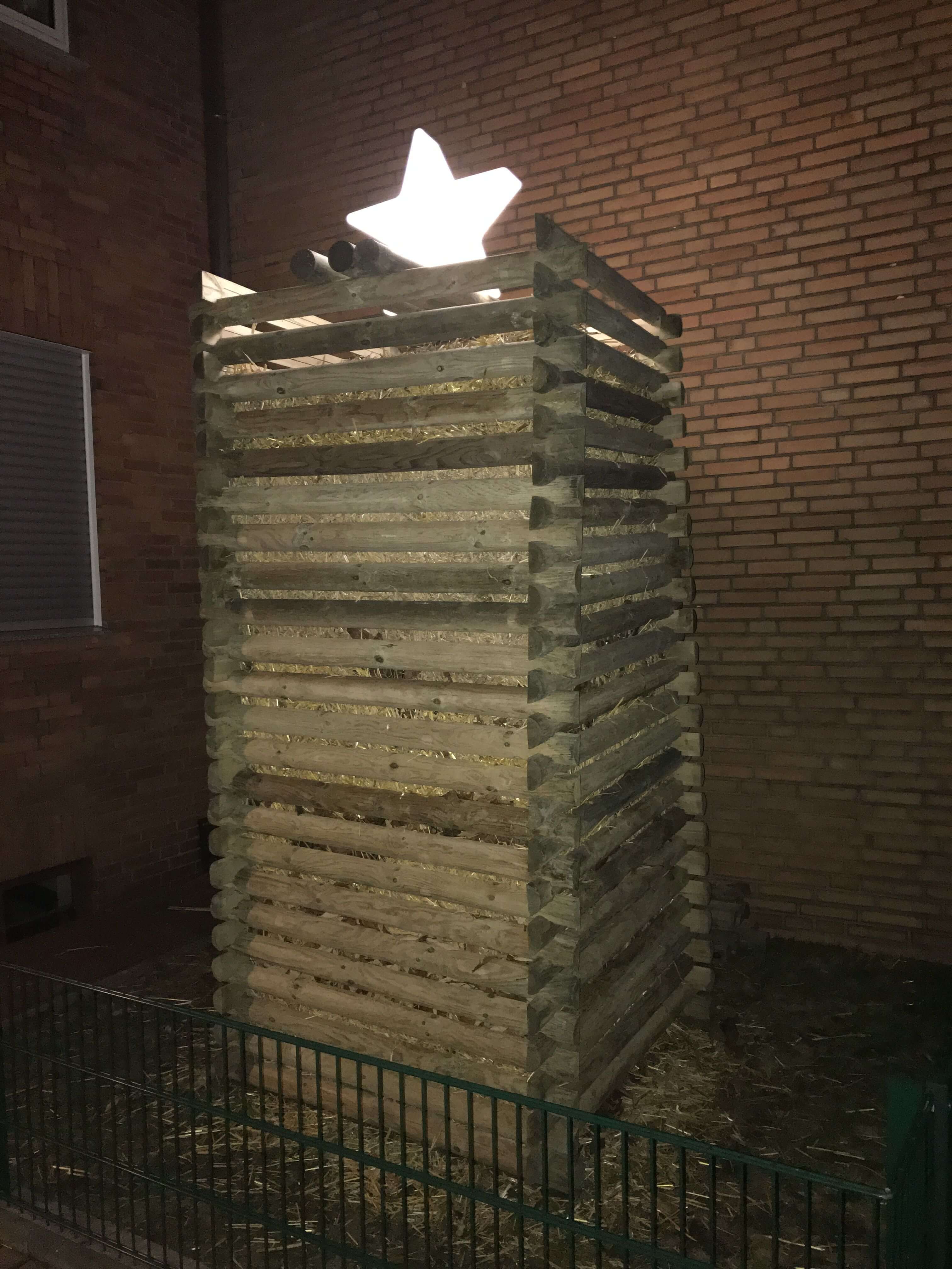 banane_stern Gestapelte Holzkonstruktion mit Strohfüllung und leuchtendem Stern oben, grüner Metallzaun im Vordergrund, rote Backsteinwand im Hintergrund bei Nacht
