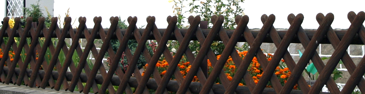 Brauner hölzerner Lattenzaun mit diagonal gekreuzten Stäben im Rautenmuster vor einem Blumenbeet mit orangefarbenen Blüten und grünen Pflanzen; im Hintergrund weiße Hauswand und niedrige Steinmauer