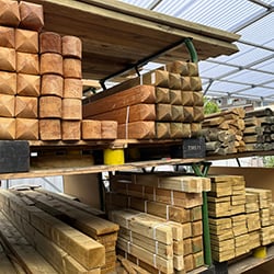 Gestapelte Bauholzware: gehobelte Balken, quadratische Pfosten und Bretter in verschiedenen Stärken auf Metallregalen unter lichtdurchlässigem Hallendach, mehrere Bündel mit Kunststoffbändern