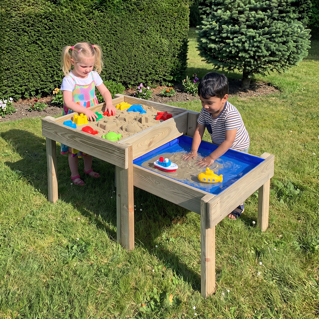 Zwei Kinder spielen an einem hölzernen Sand- und Wassertisch im Garten; Mädchen mit Zöpfen formt Sand mit Förmchen, Junge rührt im Wasser mit Spielbooten, Rasen und Hecke im Hintergrund
