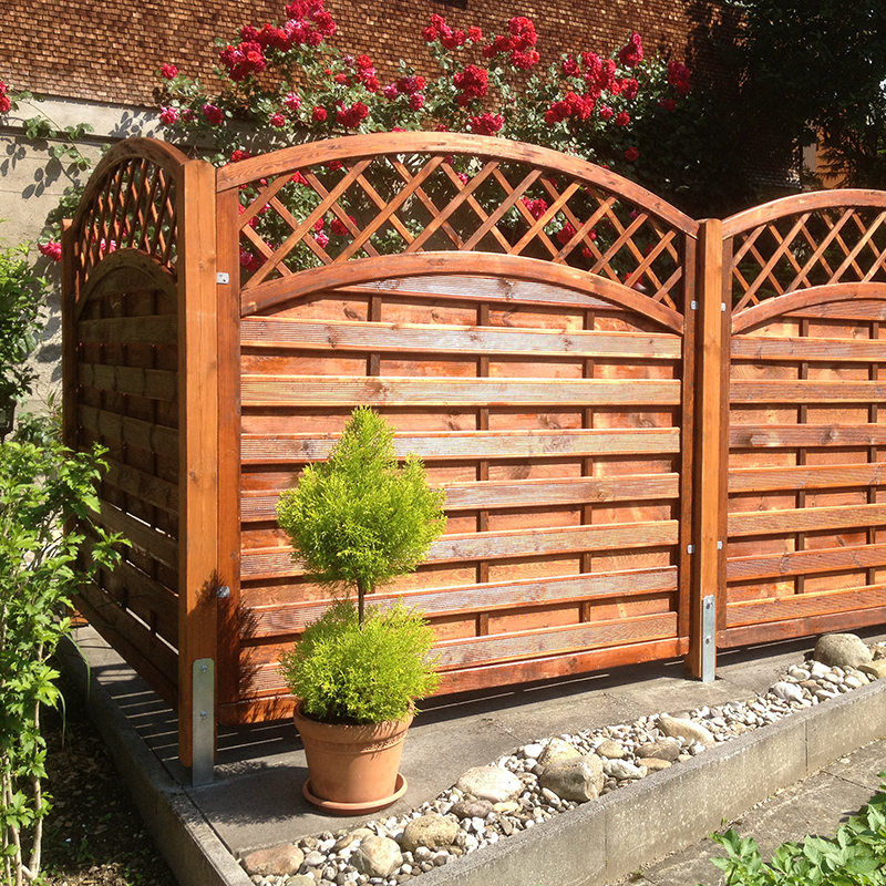 cecina_kunde_2017-07 Sonniger Garten mit geschwungenem Holzsichtschutz mit Rautenmuster, davor Terrakotta-Topf mit zweistufiger Konifere, Kies- und Steinrand und im Hintergrund blühende rote Rosen