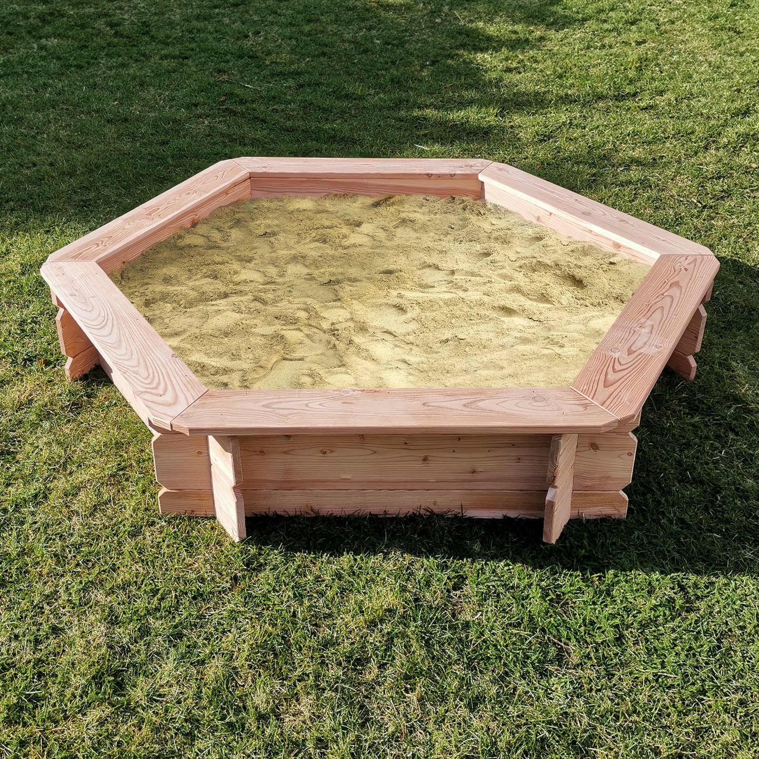 Sandkasten Lina Sechseck aus Lärchenholz, Durchmesser 174 cm, für den Garten Sechseckiger Sandkasten aus Lärchenholz mit eingebauter Sitzfläche, Modell "Lina", Durchmesser 174 cm, auf einer Rasenfläche stehend, Sand im Inneren sichtbar