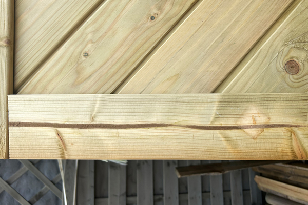 Nahaufnahme von hellen Kieferholzplanken: diagonale und horizontale Bretter, sichtbare Maserung, Knoten und Fugen, darunter unscharfe graue Holzlatten im Hintergrund