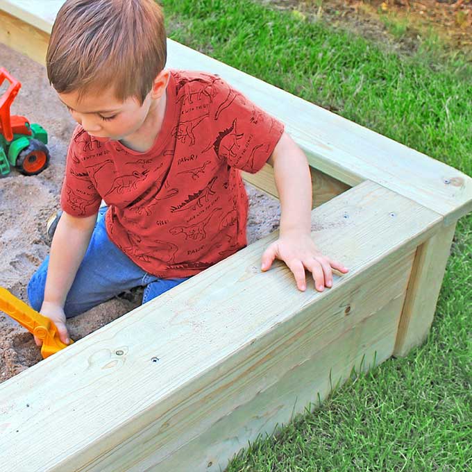 Kinderwelt Kleiner Junge sitzt im Holzsandkasten auf dem Rasen, trägt rotes Dinosaurier-T-Shirt und gräbt mit gelber Schaufel; grüner Spielzeugtraktor liegt daneben.