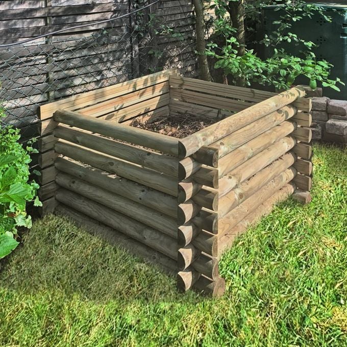 Quadratisches, erhöhtes Hochbeet aus miteinander verzahnten Rundholzstämmen auf Rasen, gefüllt mit Erde und Laub, am Holzzaun zwischen grünen Pflanzen