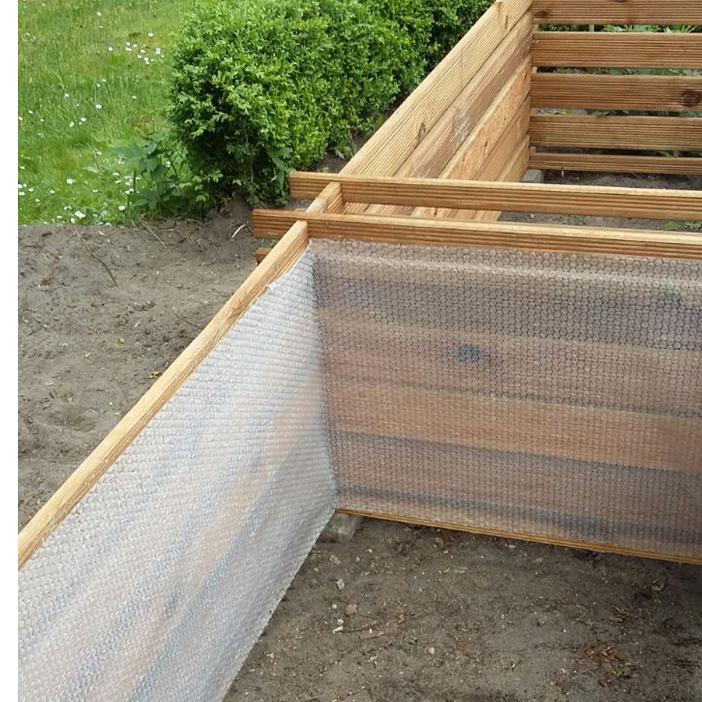 Eckansicht eines hölzernen Hochbeets im Bau mit quer verlaufenden Holzlatten und innen ausgelegter Luftpolsterfolie, umgeben von nacktem Boden; im Hintergrund Rasen und eine dichte Hecke.