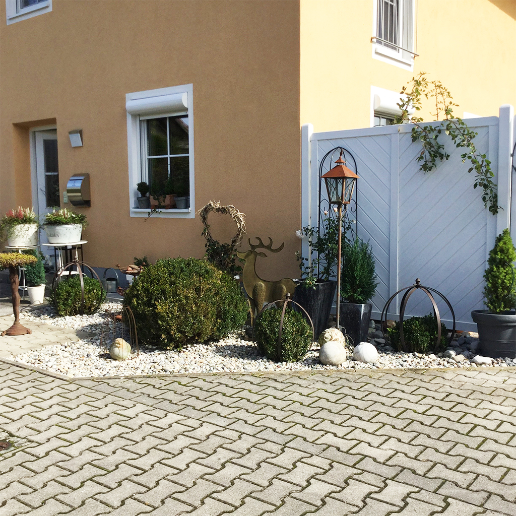 Vorgarten eines gelben Hauses mit weißem Sichtschutzzaun, Kiesbeet mit Buchsbaumkugeln, Metallhirsch-Skulptur, Laterne, Pflanzkübeln und Dekorkugeln, gepflasterte Einfahrt im Vordergrund