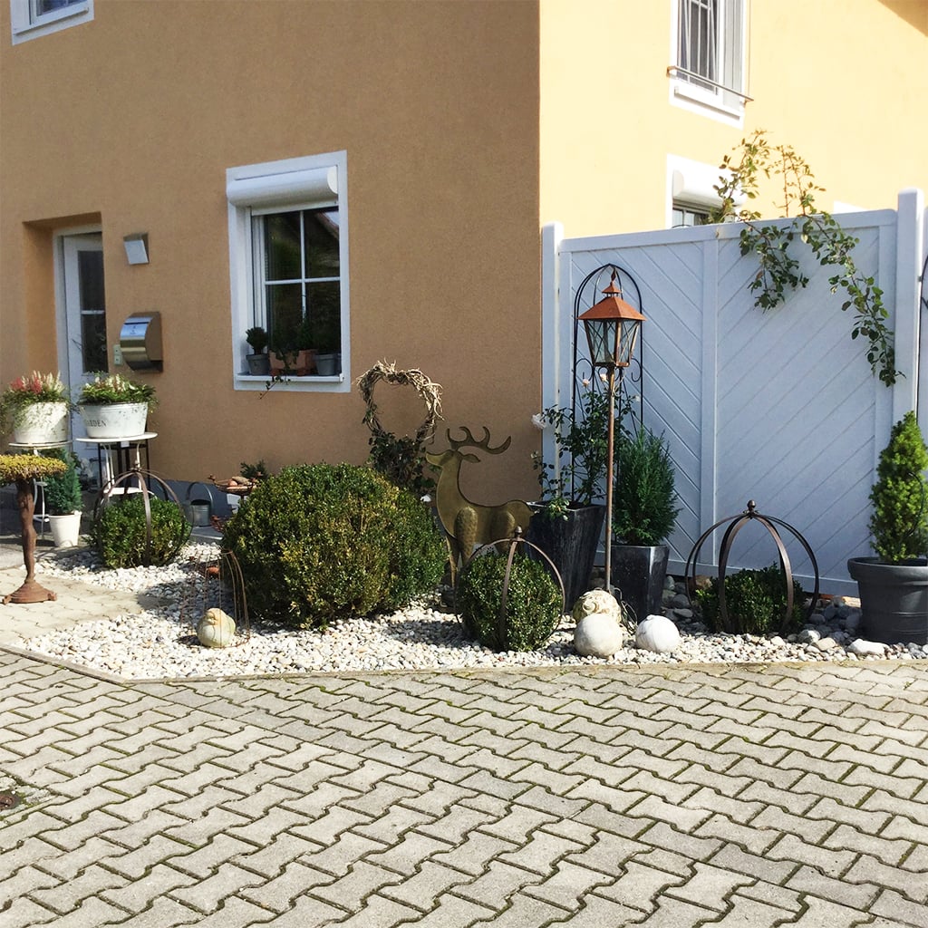 stabil-massivweiss-kundenfoto Vorgarten eines gelben Hauses mit weißem Sichtschutzzaun, Kiesbeet mit Buchsbaumkugeln, Metallhirsch-Skulptur, Laterne, Pflanzkübeln und Dekorkugeln, gepflasterte Einfahrt im Vordergrund