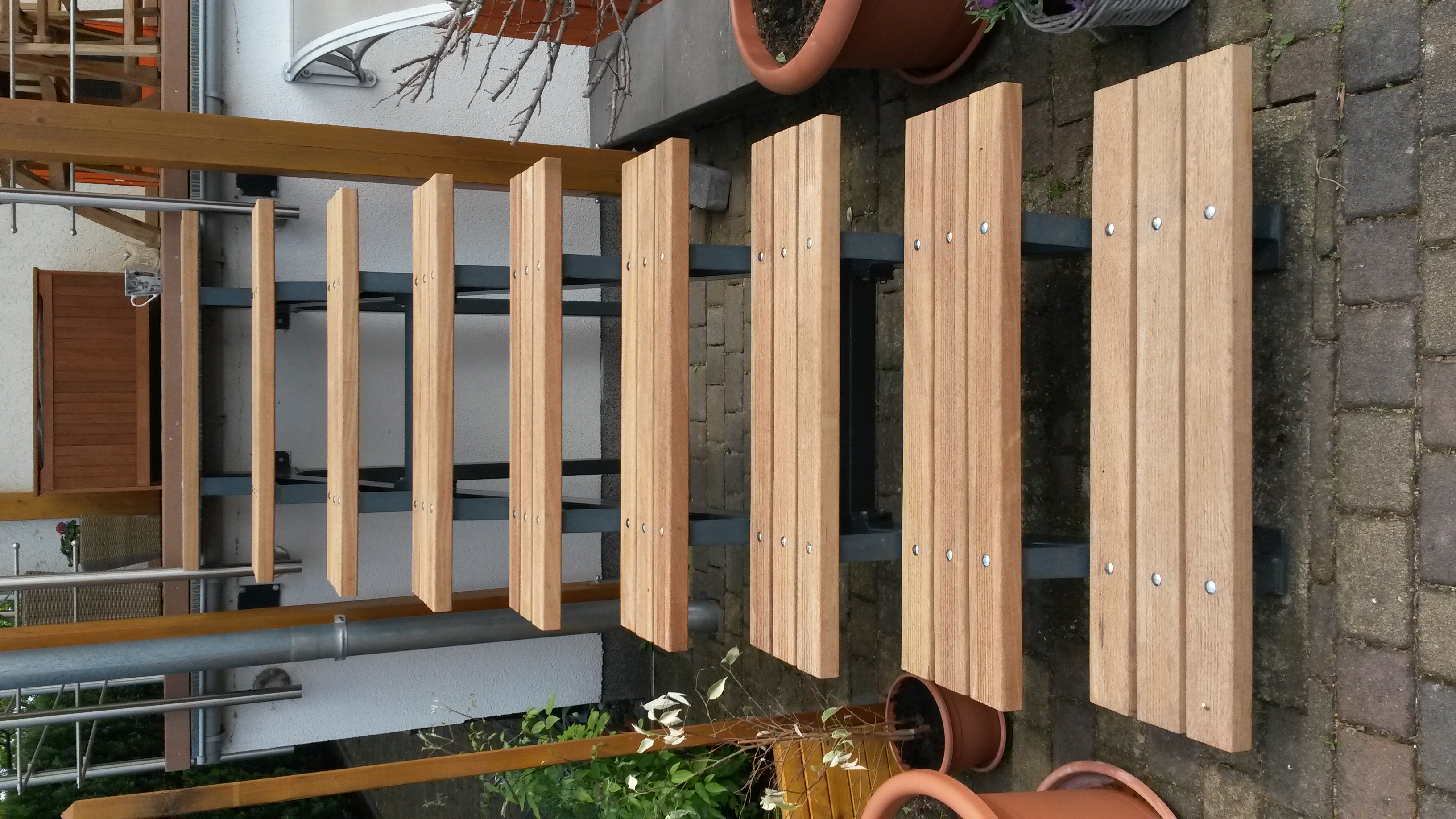 Offene Außentreppe mit mehreren hellen Holzstufen auf grauen Metallträgern, führt zu einer kleinen Terrasse; links Regenrohr und Pflanzenkübel, rechts Topfpflanzen und kahle Äste, oberhalb Holzbrüstung und Tasse auf der Veranda