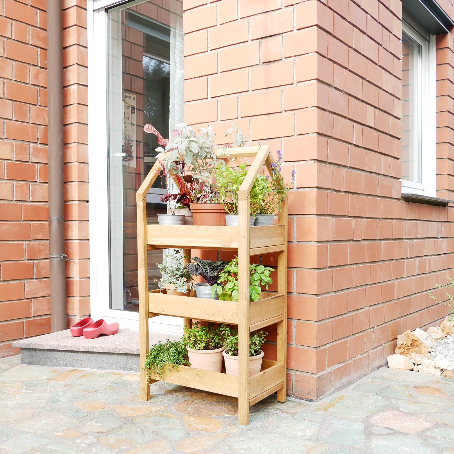 Gartenregal Ares aus hellbraun imprägnierter Fichte mit verschiedenen Pflanztöpfen auf drei Ebenen auf einer Terrasse Gartenregal Ares aus hellbraun imprägnierter Fichte mit drei Ablagen, auf denen verschiedene Blumentöpfe mit Pflanzen stehen, platziert auf einer Terrasse vor einer Hauswand aus roten Ziegeln