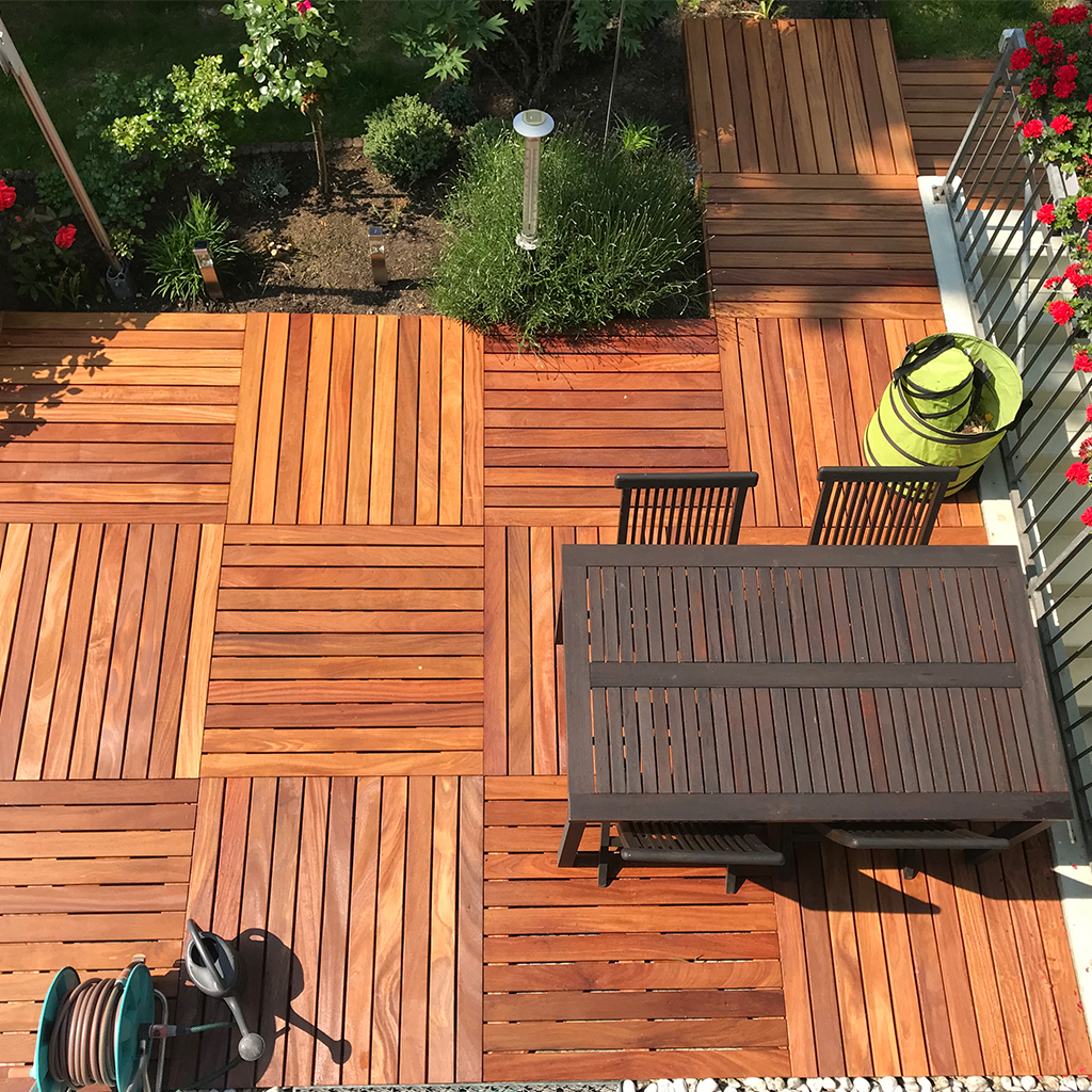cumaru-holzfliese100-2 Draufsicht auf Terrasse mit quadratischen Holzfliesen in wechselnder Maserung, dunkler Holztisch und zwei Stühlen, rechts Balkon mit roten Blumen, oben Beet mit Sträuchern und Laterne, unten Gartenschlauch auf Rolle