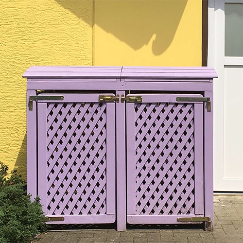 Muelltonnenbox_Viol_Flieder_500x500 Lavendelfarbene hölzerne Doppeltür mit diagonaler Lattenrasterung und Metallbeschlägen vor gelber Hauswand neben weißer Tür auf gepflastertem Boden