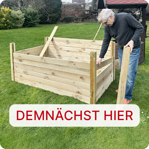 Älterer Mann baut im Garten ein rechteckiges Hochbeet aus Holzlatten auf grünem Rasen; im Vordergrund ein weißes Schild mit roter Aufschrift DEMNÄCHST HIER