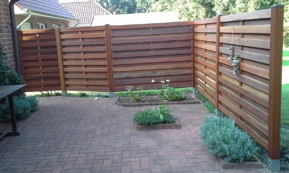 Gepflasterte Terrasse mit horizontalem Holz-Sichtschutzzaun an drei Seiten, zwei kleinen erhöhten Beeten mit vereinzelten Pflanzen und hängender Steindekoration am rechten Zaunpfosten
