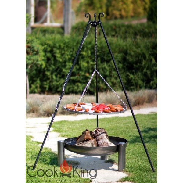 Dreibein-Grill mit hängendem Grillrost über Feuerschale; Rost belegt mit Würstchen und Gemüse, darunter Holzscheite, im Garten auf Grasflächen