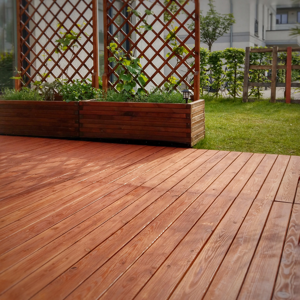 Holzterrasse im Vordergrund, links zwei erhöhte Holzpflanzkästen mit Rankgittern und Kletterpflanzen, im Hintergrund Rasen, Hecke und Wohngebäude