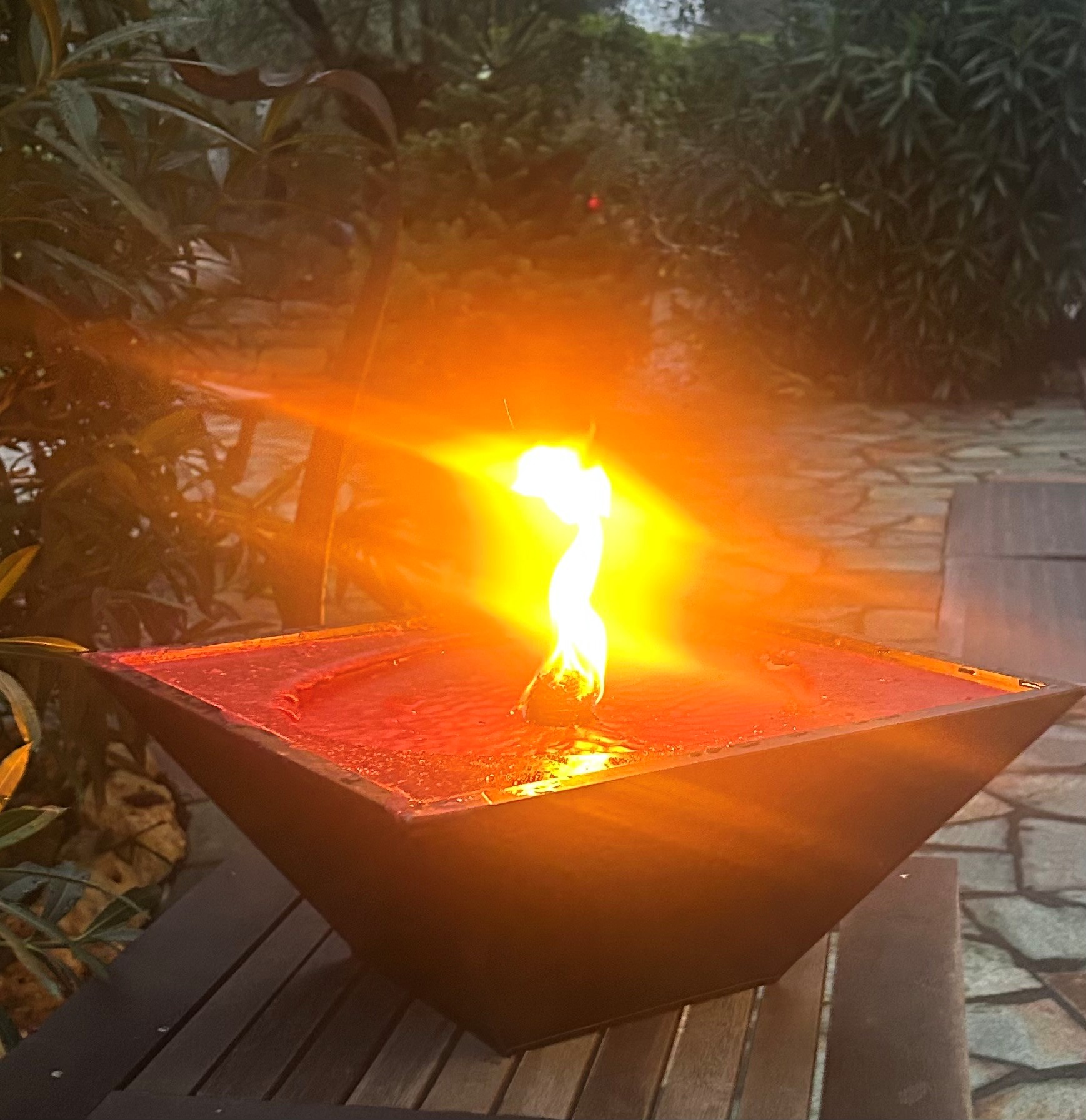 Rote Wachsschale mit brennender Flamme im Garten Rote Wachsschale mit brennender Flamme in einem schwarzen, geometrischen Behälter im Freien auf einem Holztisch, umgeben von Pflanzen und Steinboden.