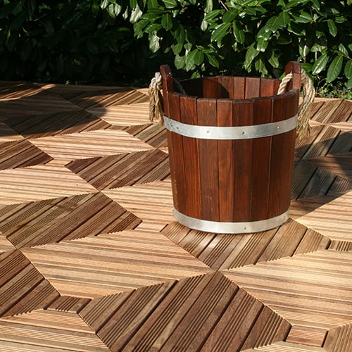 Cumaru_diagonal_fliese-500x500 Holzeimer mit Metallbändern und Taugriffen auf geometrisch gemusterten Holzfliesen-Terrasse, teilweise im Sonnenlicht, grüner Busch im Hintergrund.